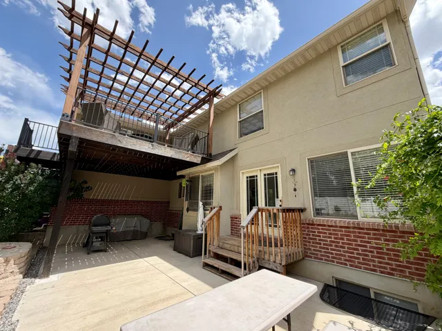 Exterior featuring a second-story deck with a wood pergola, a ground-level concrete patio, stucco siding, and a brick foundation