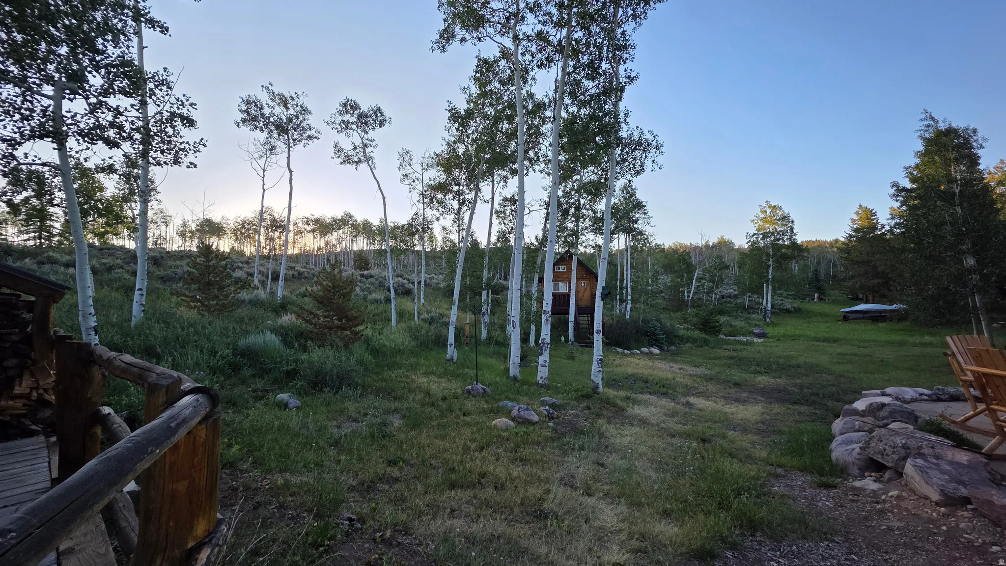 Expansive natural landscape featuring a dense forest of aspen trees, a distant cabin, and an outdoor rock-lined gathering area