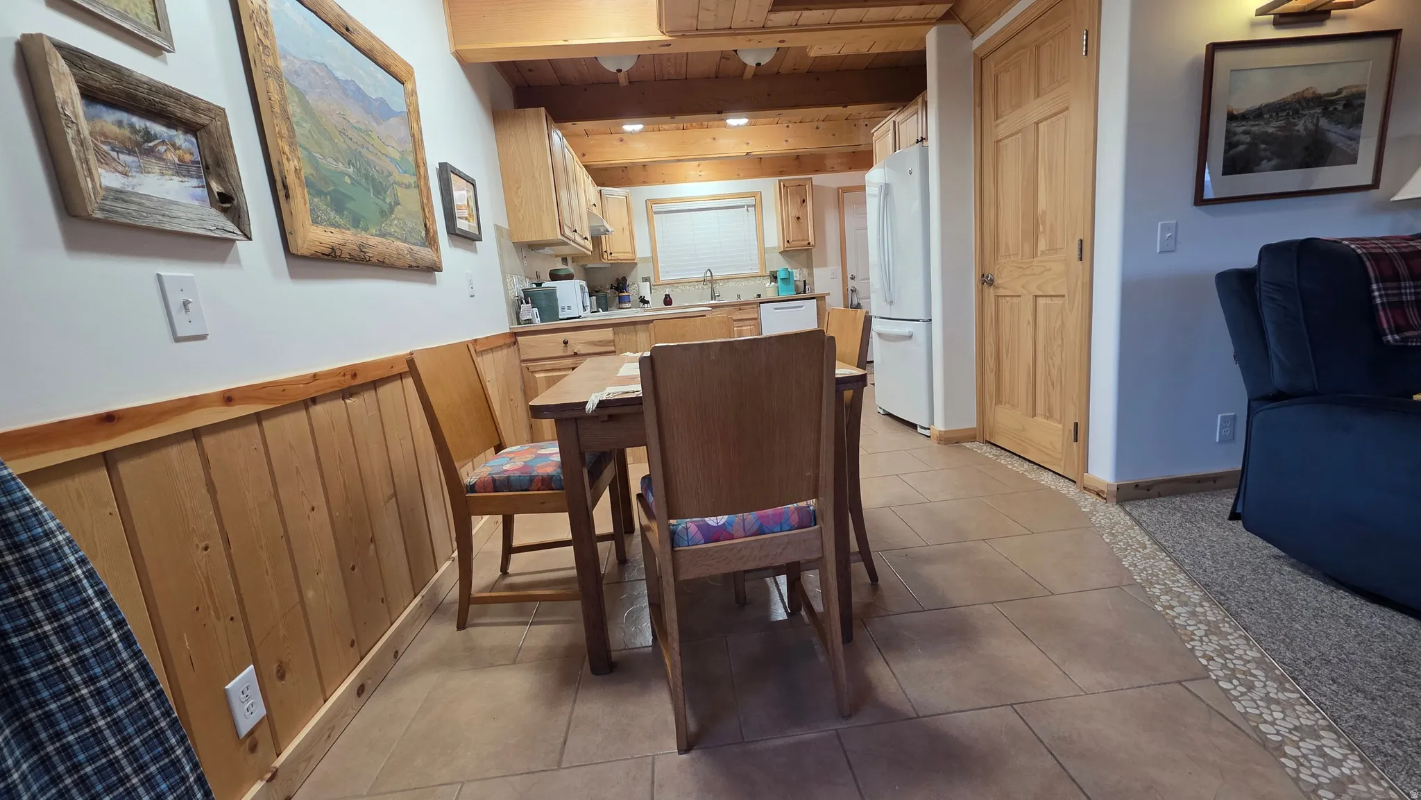 Kitchen and dining area featuring warm wood cabinetry, exposed wood beam ceiling, tile flooring, and a white refrigerator