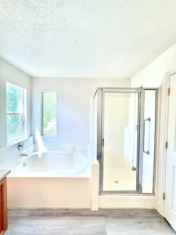 Bathroom with a built-in tub, separate glass-enclosed shower, wood-finish flooring, and two windows providing natural light