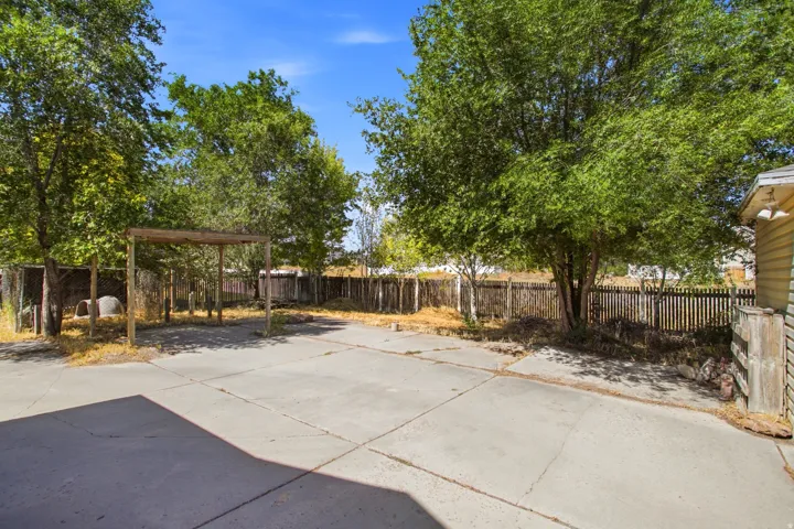 Expansive concrete patio area featuring a wooden pergola structure, mature trees, and a perimeter wooden fence