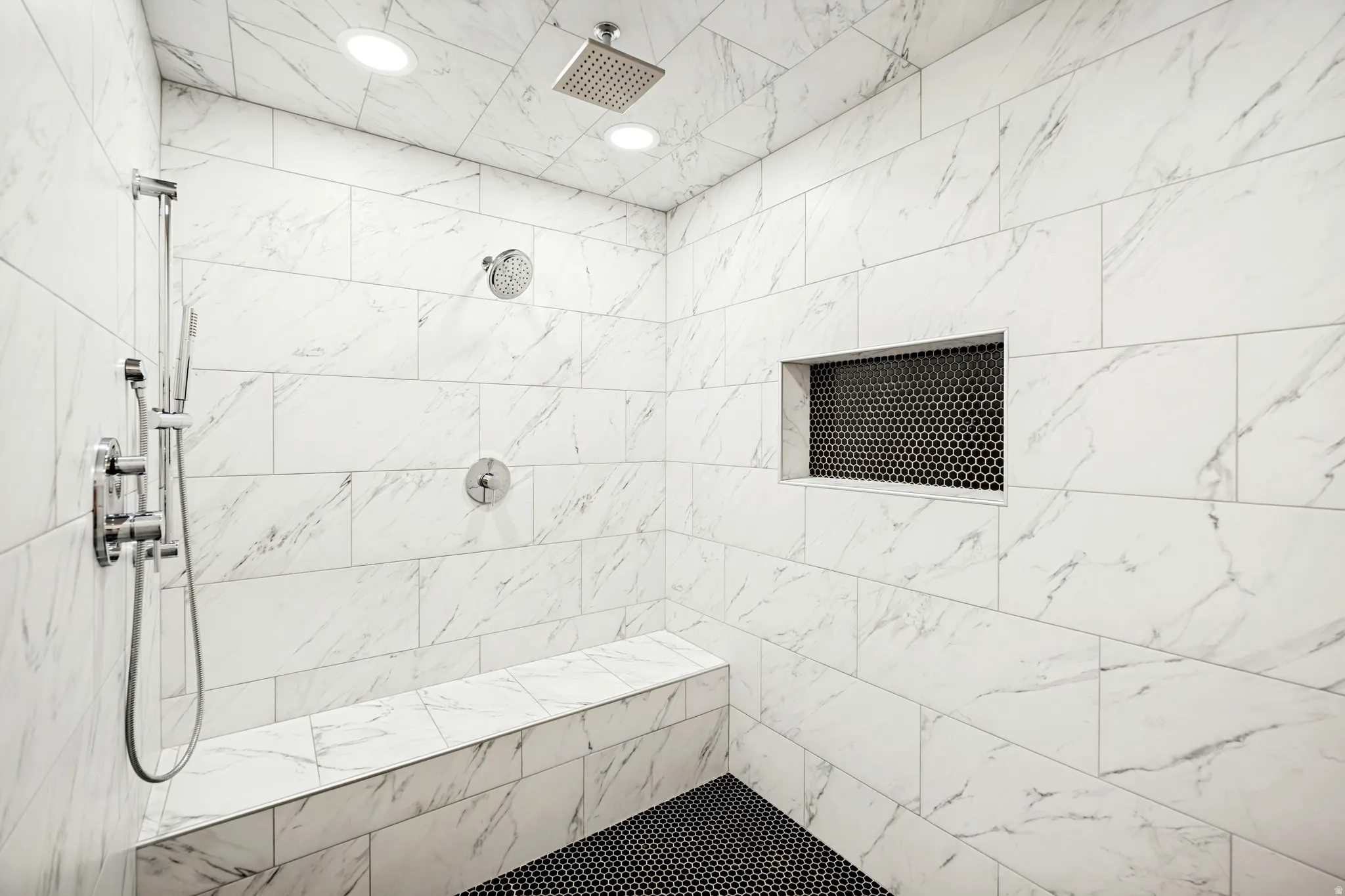 Spacious shower with large format white tile walls, a built-in bench, and black hexagon tile flooring