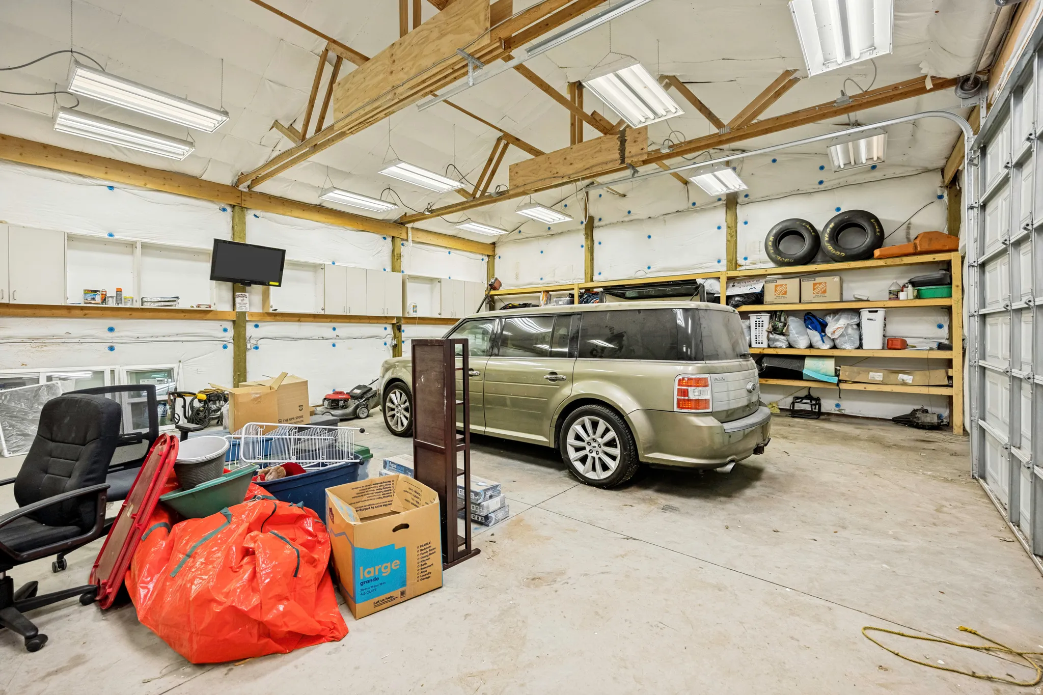 Spacious garage featuring a concrete floor, exposed wood beam ceiling, multiple fluorescent light fixtures, overhead cabinetry, and built-in shelving
