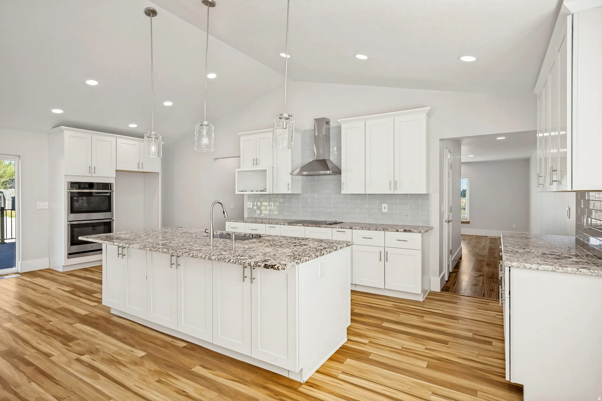 Open-concept kitchen featuring an expansive center island with an undermount sink, granite countertops, white shaker cabinetry, stainless steel double wall ovens, and wood-finish flooring
