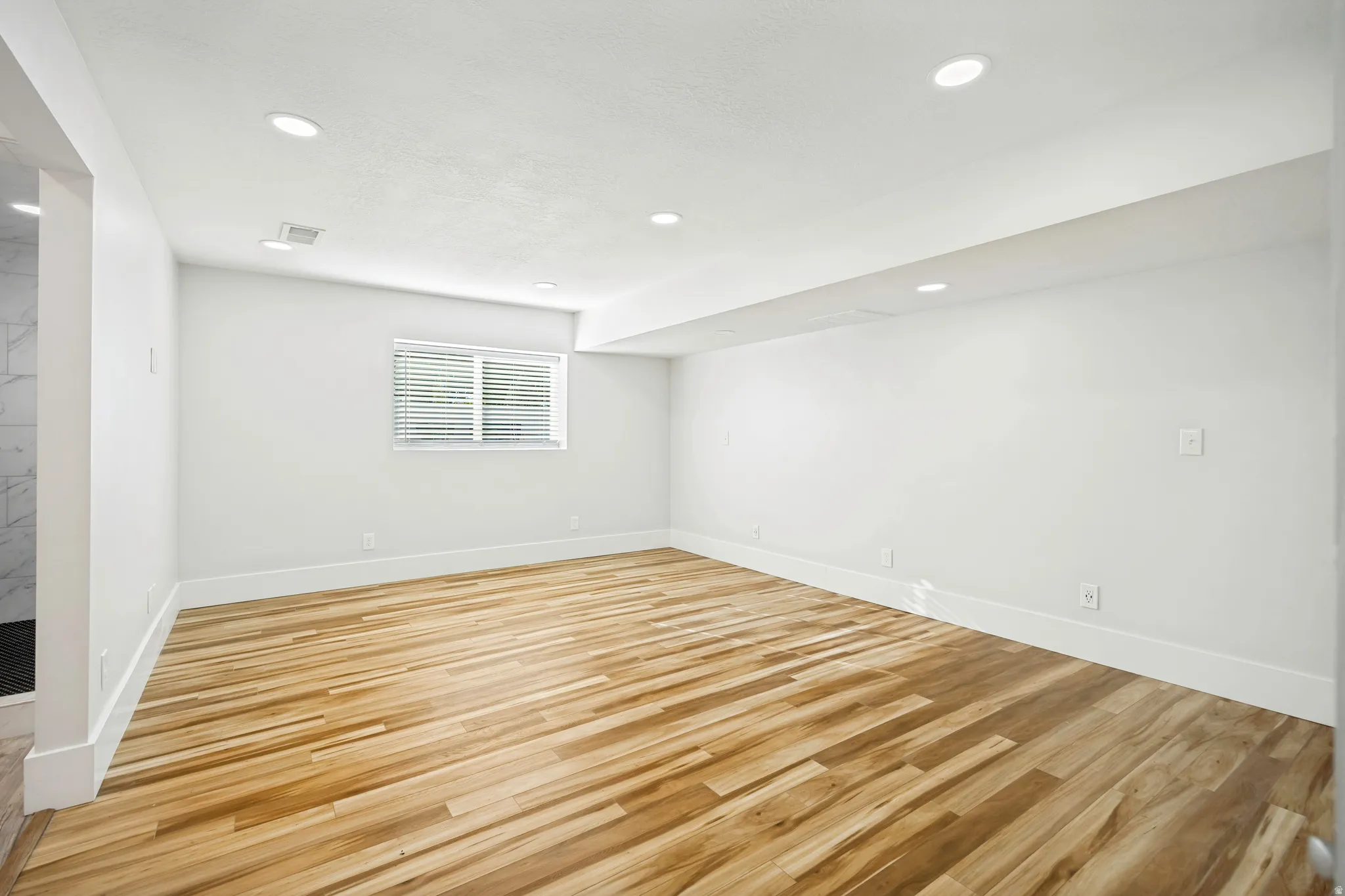 Spacious room featuring wood-finish flooring, recessed lighting, a window with blinds, crisp white walls, and substantial white baseboards