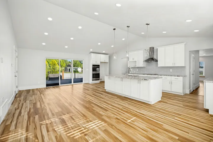Open-concept living space featuring wood-finish flooring, a contemporary kitchen, white cabinetry, a large island with a sink, and stainless steel appliances including a range hood and double wall ovens