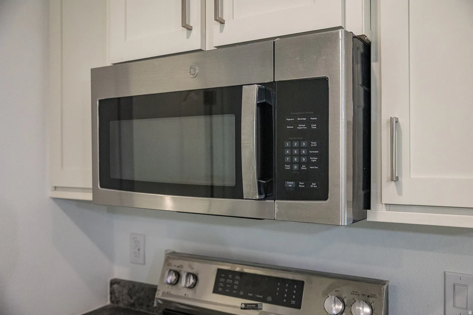Stainless steel over-the-range microwave with white shaker-style cabinetry, dark speckled countertop, and stainless steel range