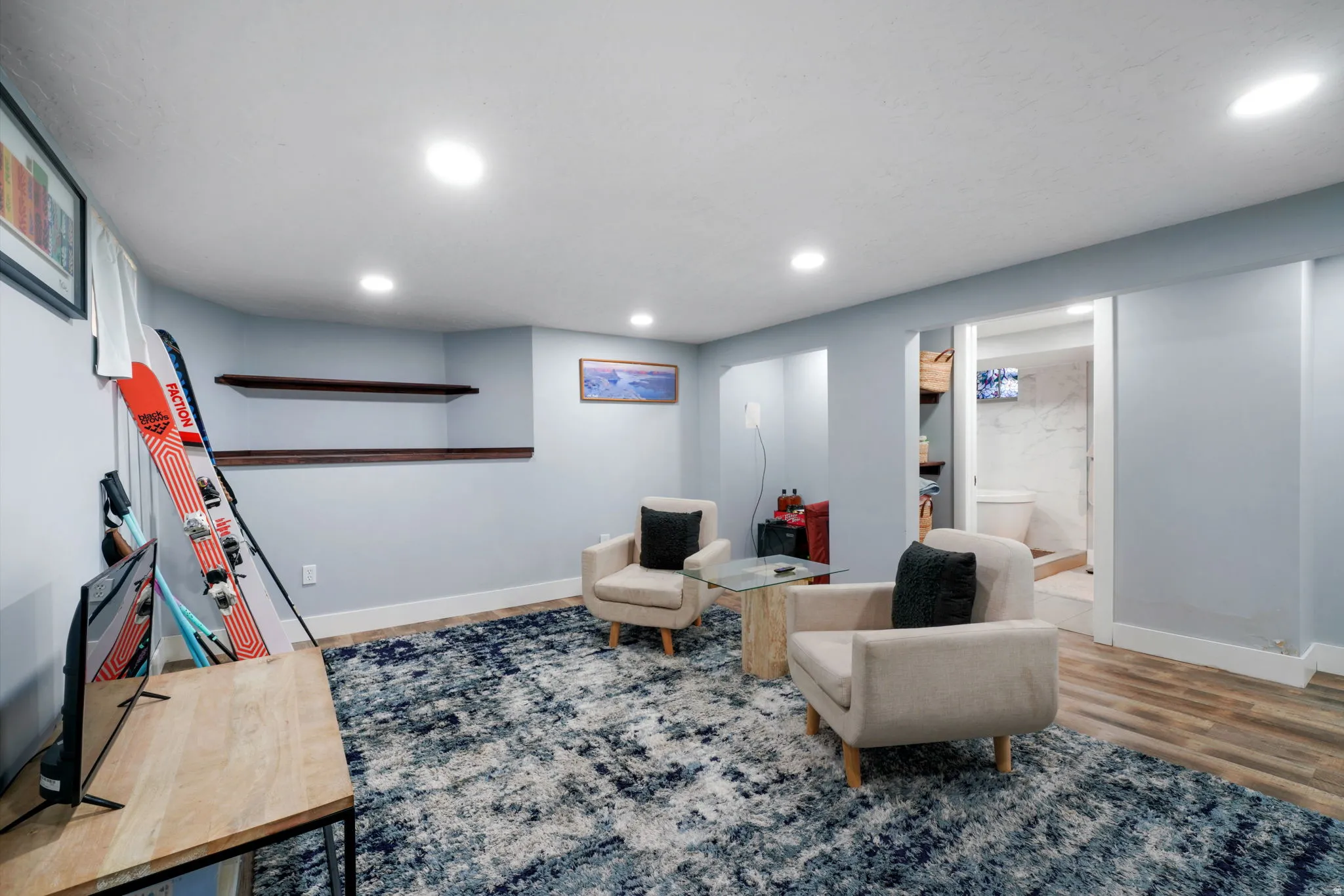 Recessed lighting illuminates this versatile space, featuring wood-finish flooring at the entrance, light blue wall tones, and two built-in wall shelves