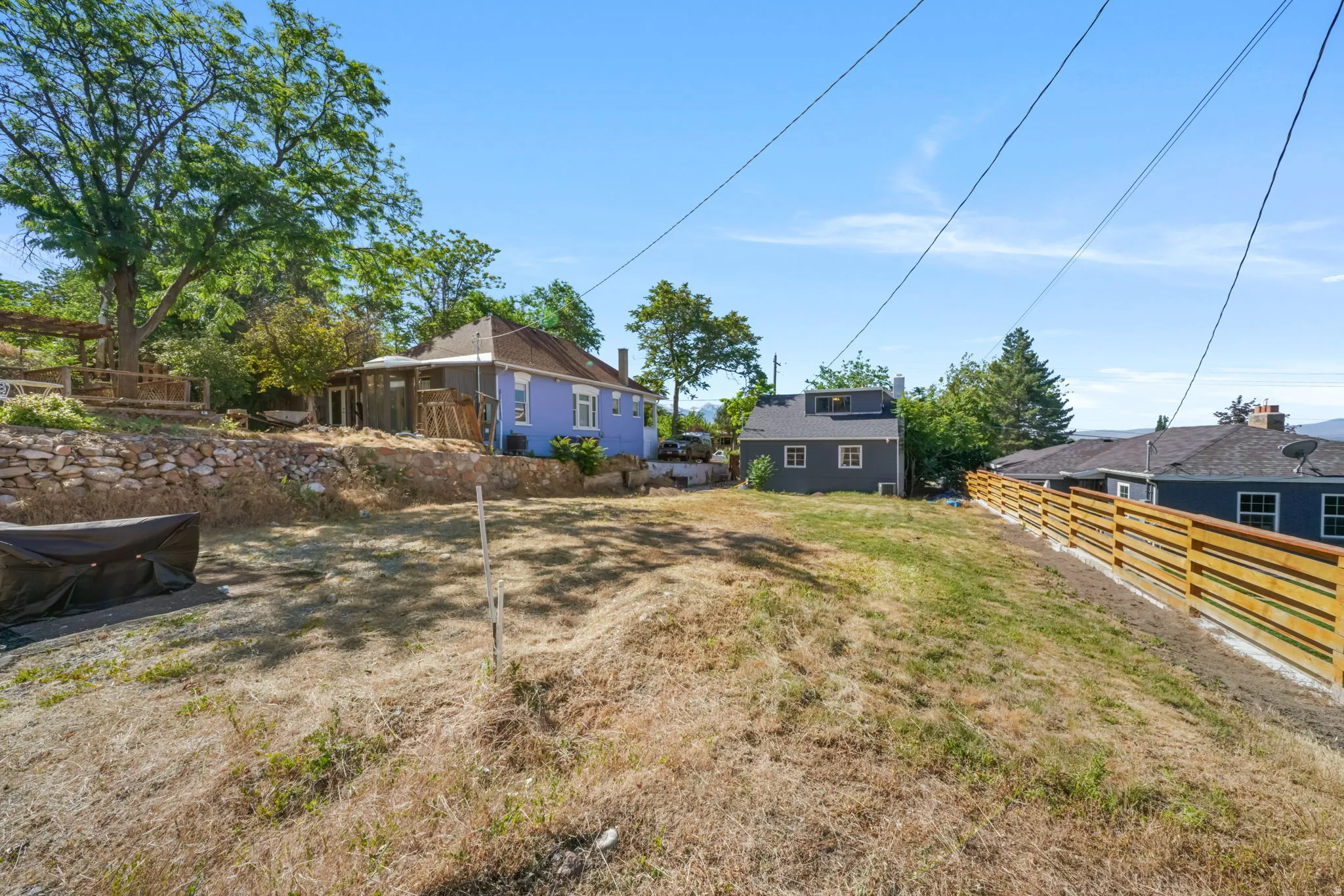 Expansive sloped lot featuring a wooden privacy fence, mature trees, and a stone retaining wall