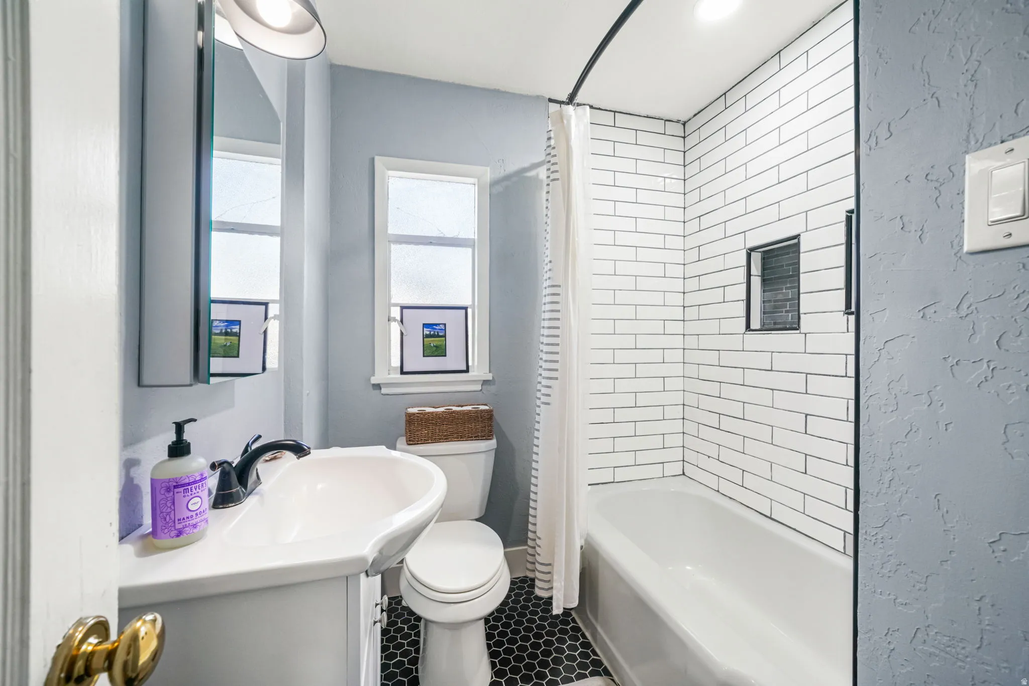 Bathroom featuring a white vanity with an integrated sink, black hexagon floor tiling, a white toilet, and a bathtub with white subway tile surround and a recessed niche