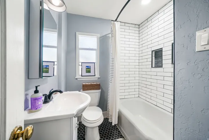 Bathroom featuring a white vanity with an integrated sink, black hexagon floor tiling, a white toilet, and a bathtub with white subway tile surround and a recessed niche