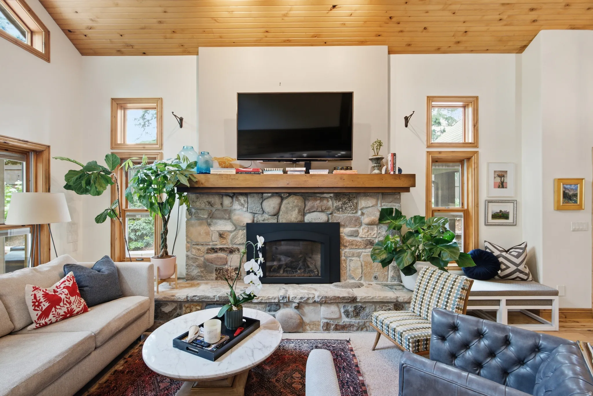 Stone-clad fireplace with a wood mantle, wood-paneled ceiling, and multiple wood-framed windows