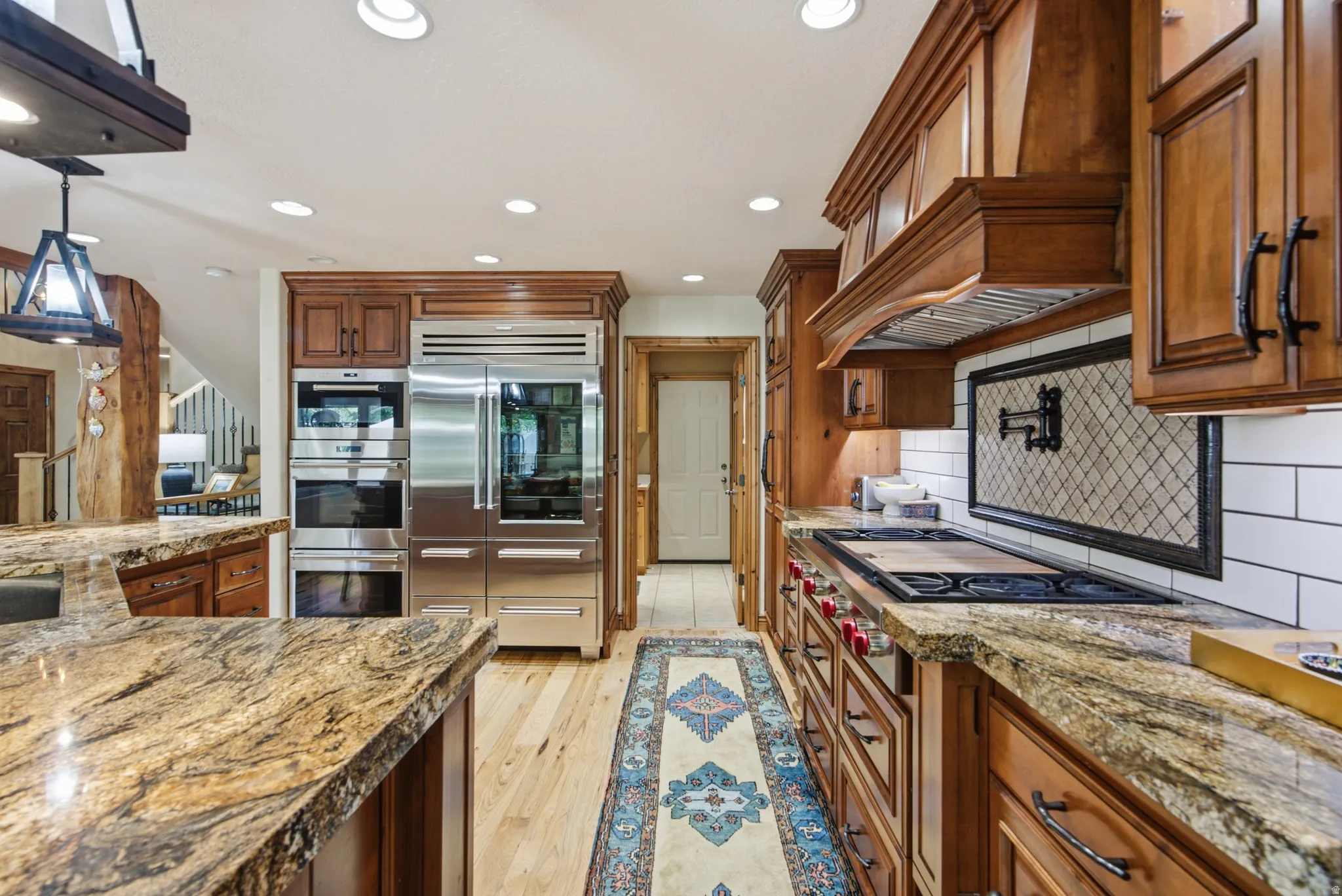 Gourmet kitchen featuring granite countertops, professional-grade stainless steel appliances, custom wood cabinetry, a tiled backsplash with decorative insert, and recessed lighting