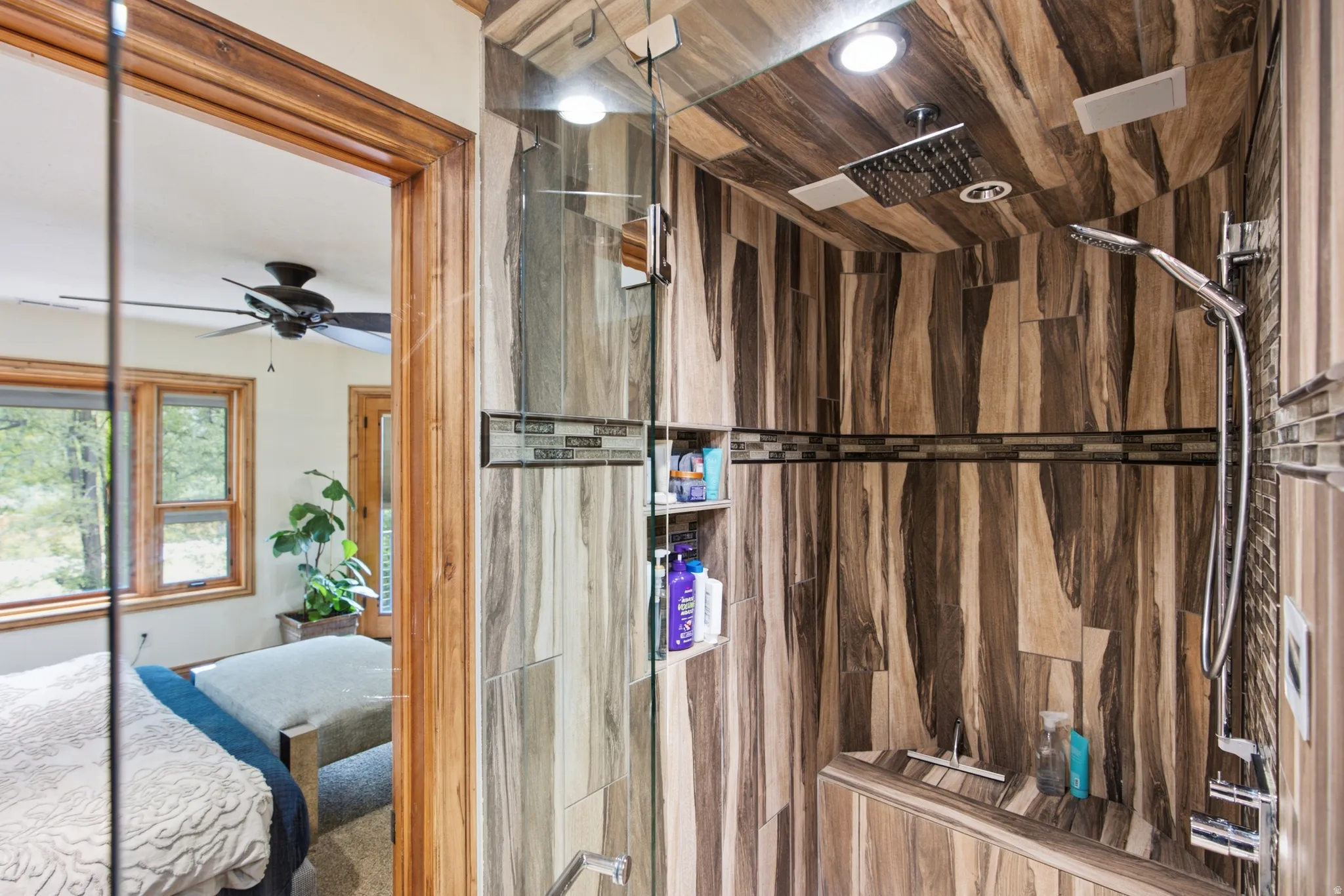 Walk-in shower featuring glass enclosure, wood-finish tile walls and ceiling, built-in recessed shelving, rain shower head, and an additional handheld shower fixture