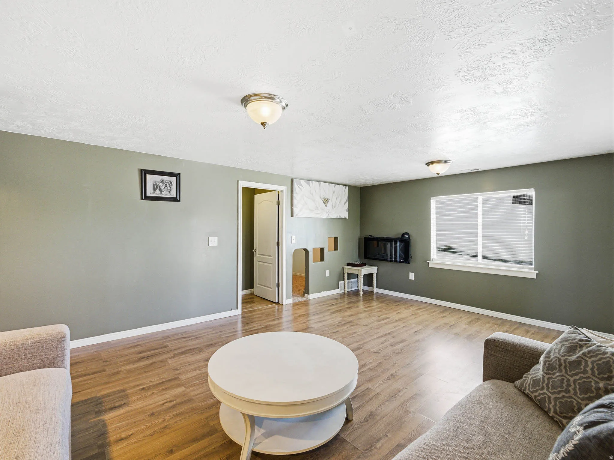 Spacious living area featuring wood-finish flooring, a neutral wall color, and a large window with blinds