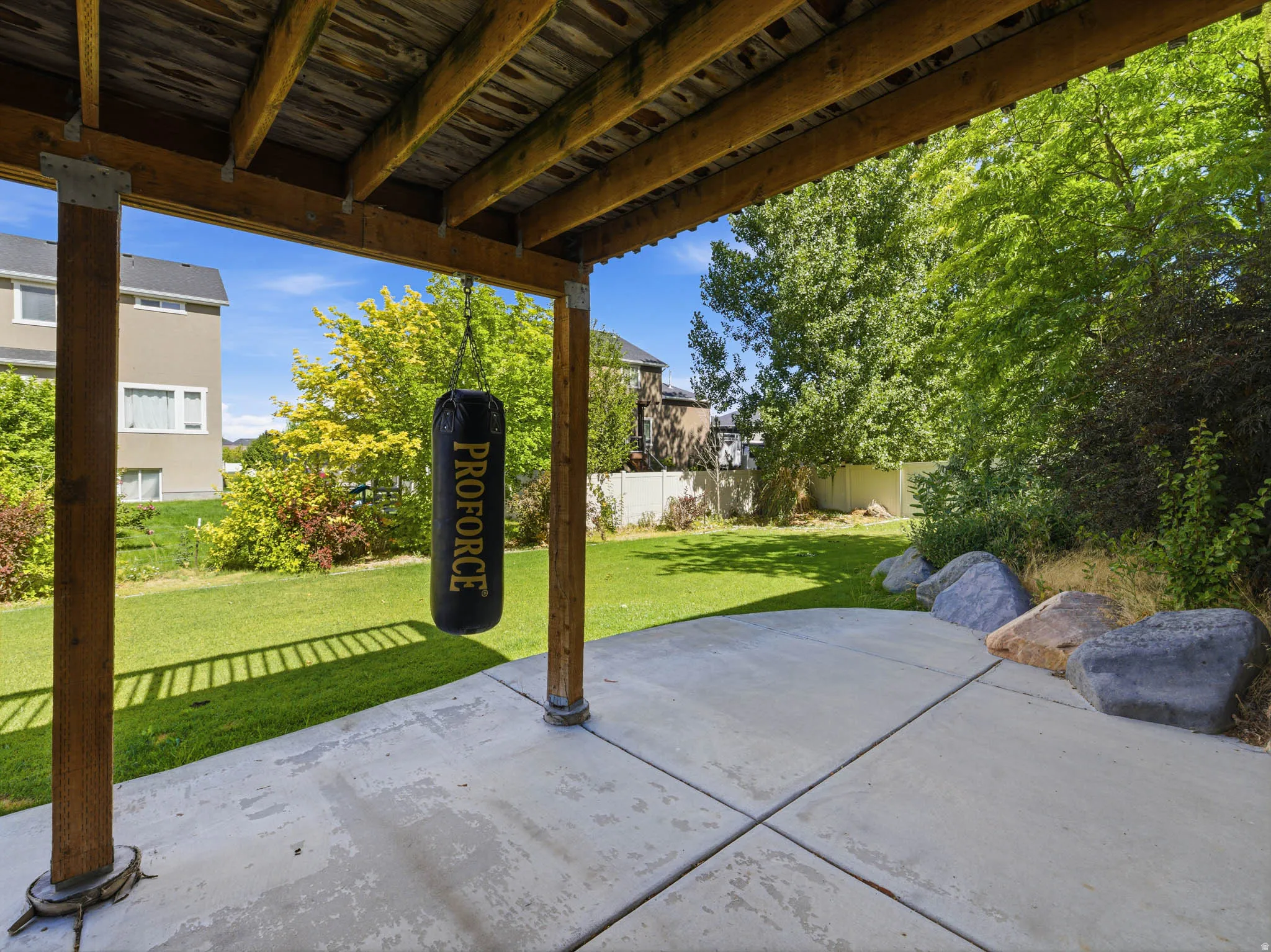 Covered concrete patio with exposed wooden beam ceiling, supported by wood posts, and featuring a verdant lawn beyond