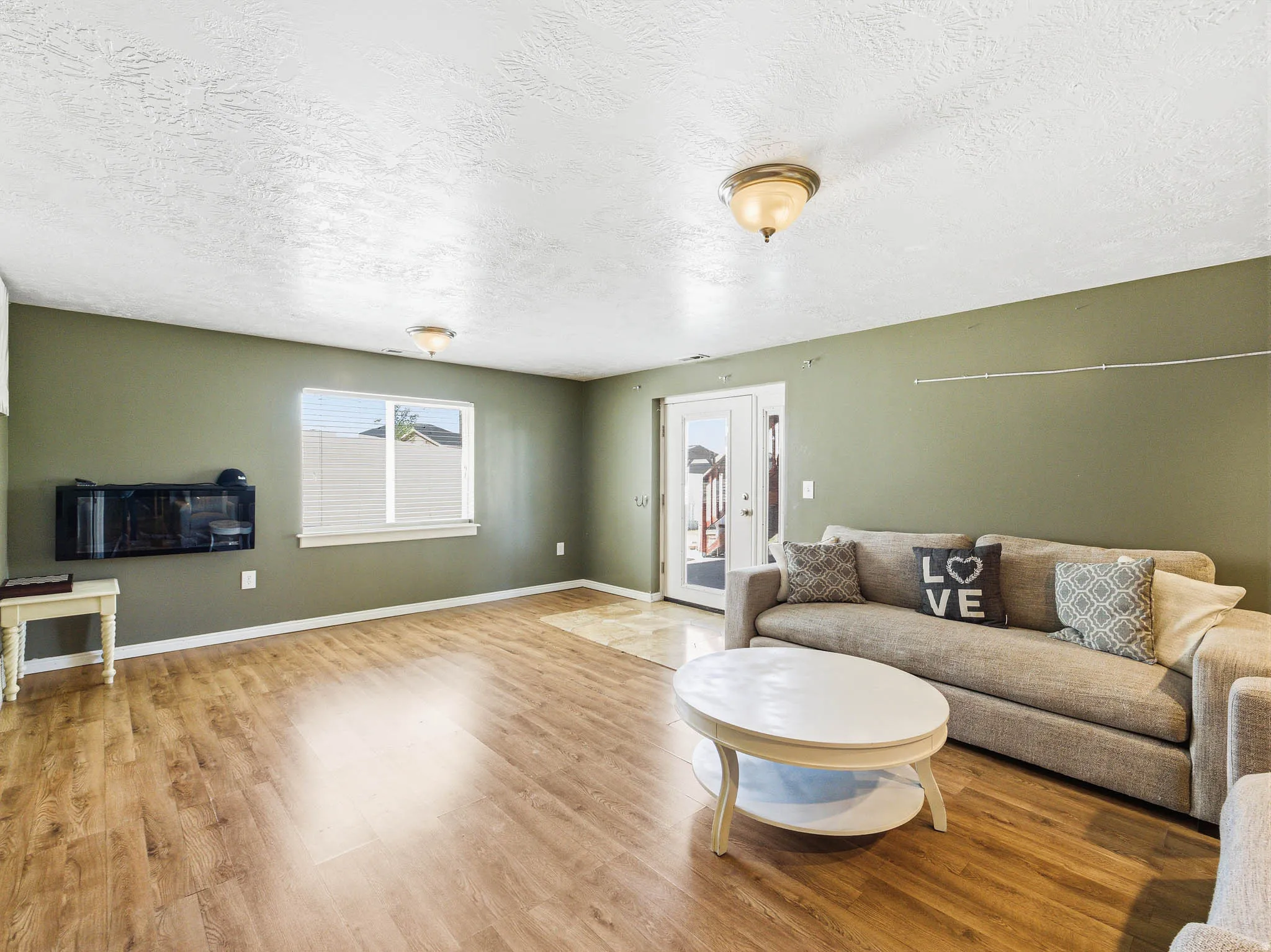 Spacious living area featuring wood-finish flooring, a contemporary wall-mounted electric fireplace, and a window with horizontal blinds