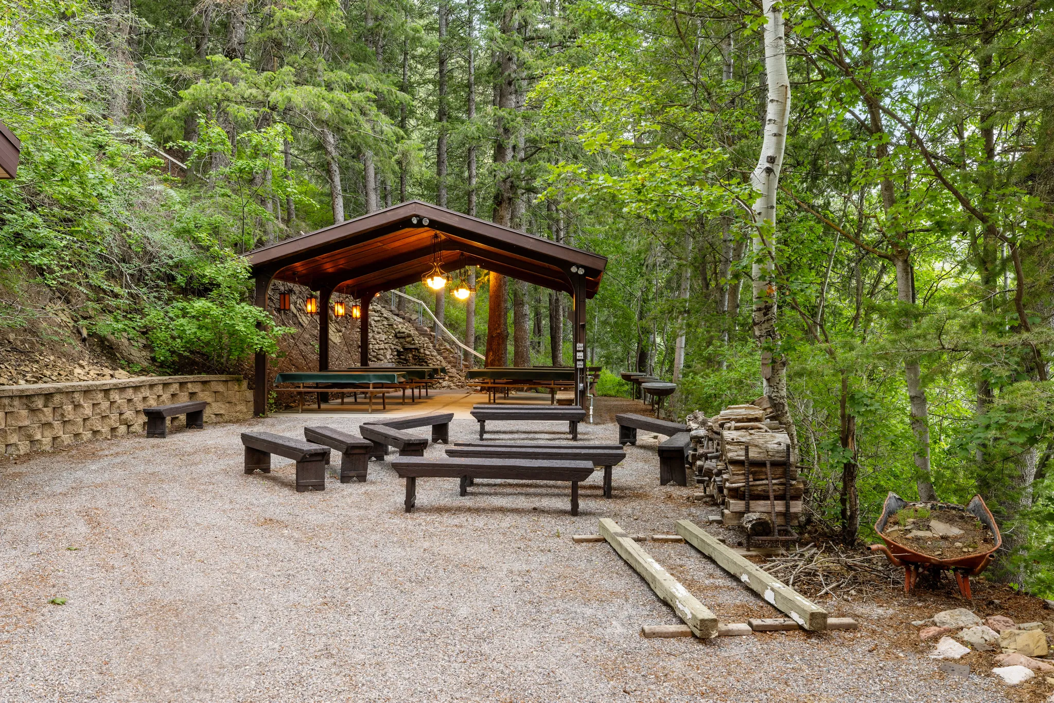 Gravel patio with a covered outdoor structure featuring pendant lighting, integrated seating, and surrounding mature trees