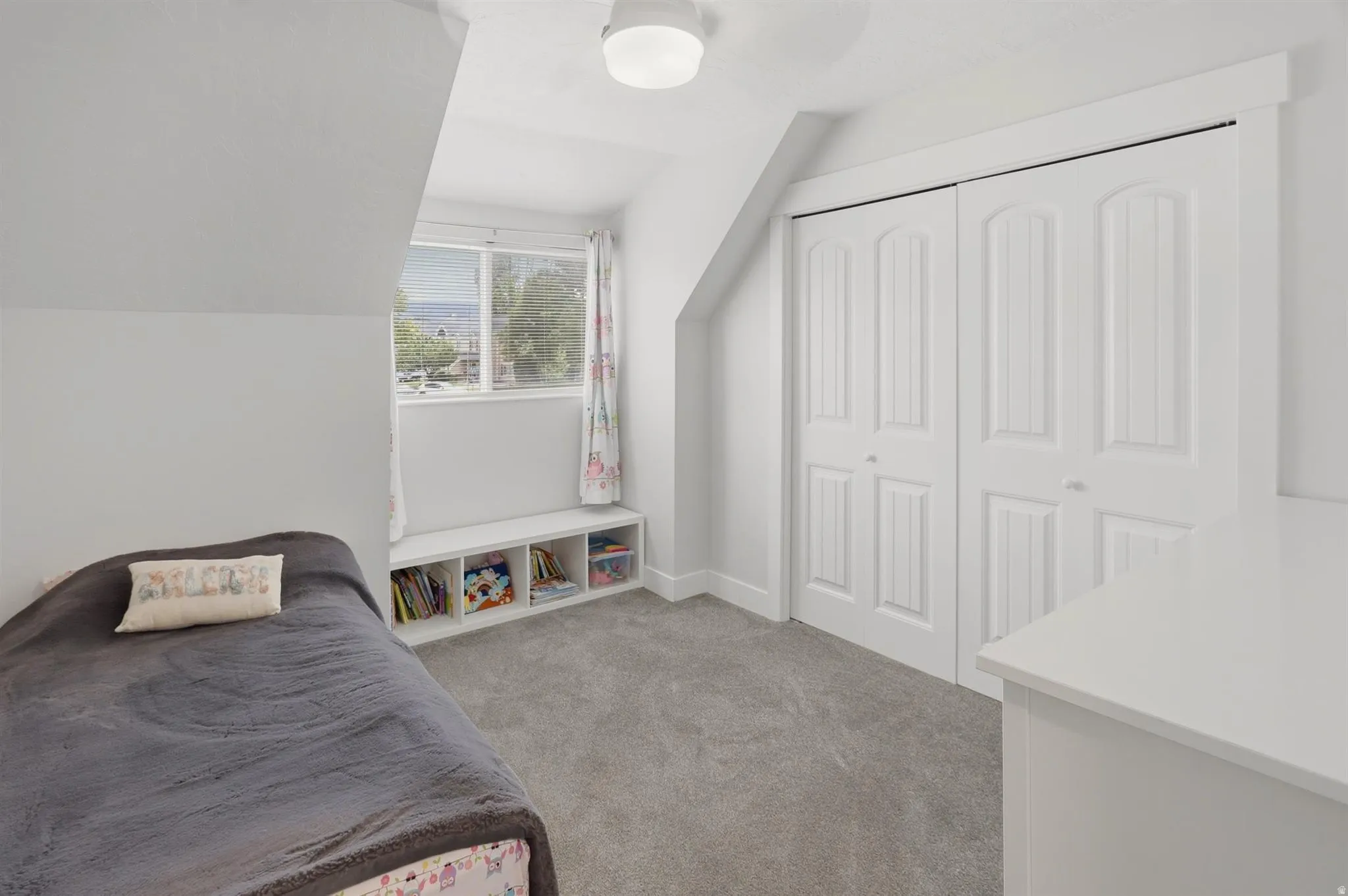 Room featuring light gray carpeting, crisp white walls, a built-in window bench with open storage, a window with exterior views, and bi-fold closet doors