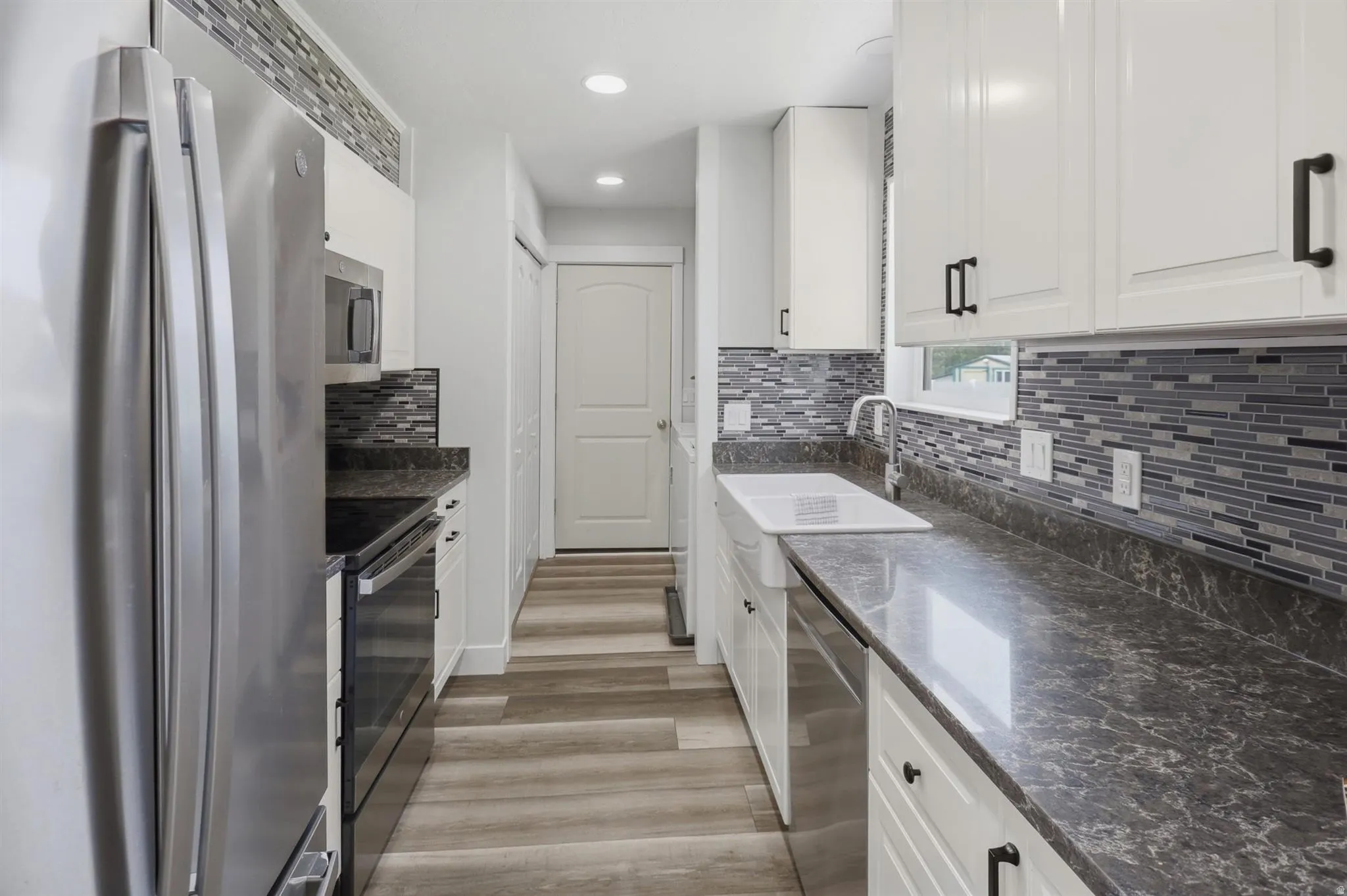 Galley kitchen featuring wood-finish flooring, white cabinetry with dark hardware, dark countertops, and a tiled backsplash