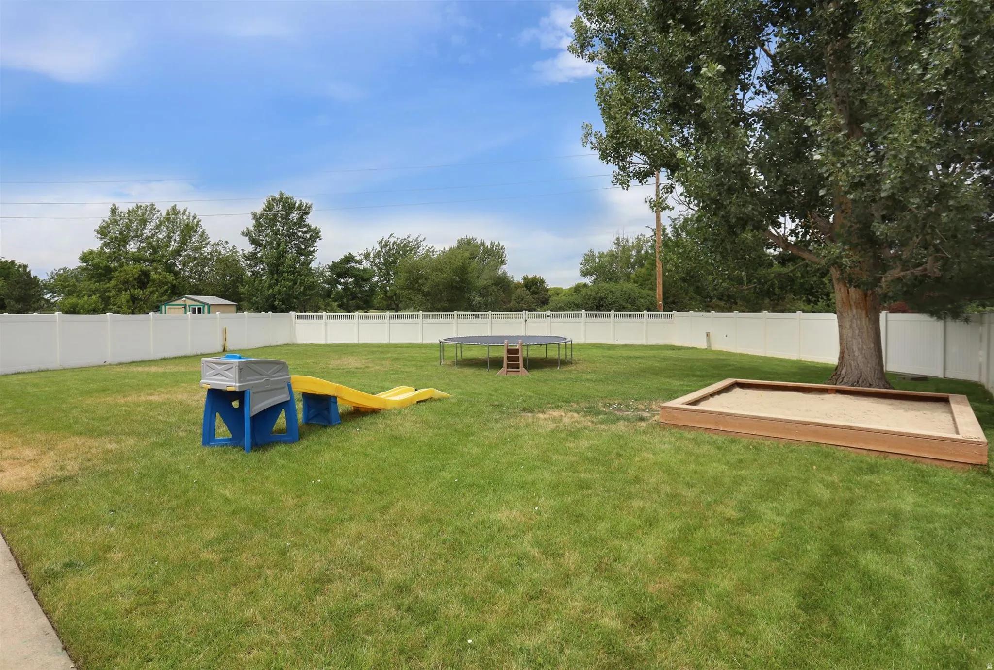 Expansive backyard featuring a manicured lawn, complete white vinyl fencing, a large mature tree, and a built-in sandbox