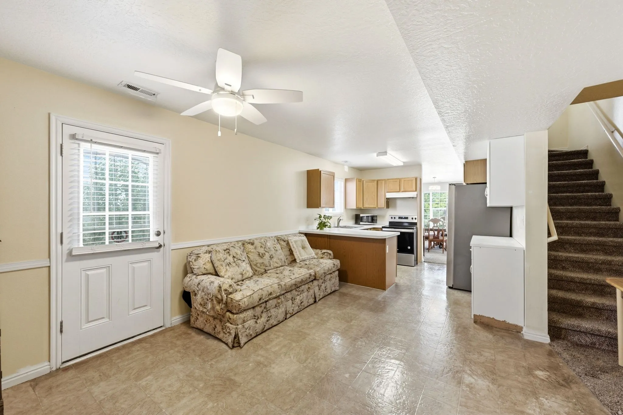 Open concept living area with light-toned flooring, featuring a kitchen space with wood cabinetry and a stainless steel refrigerator