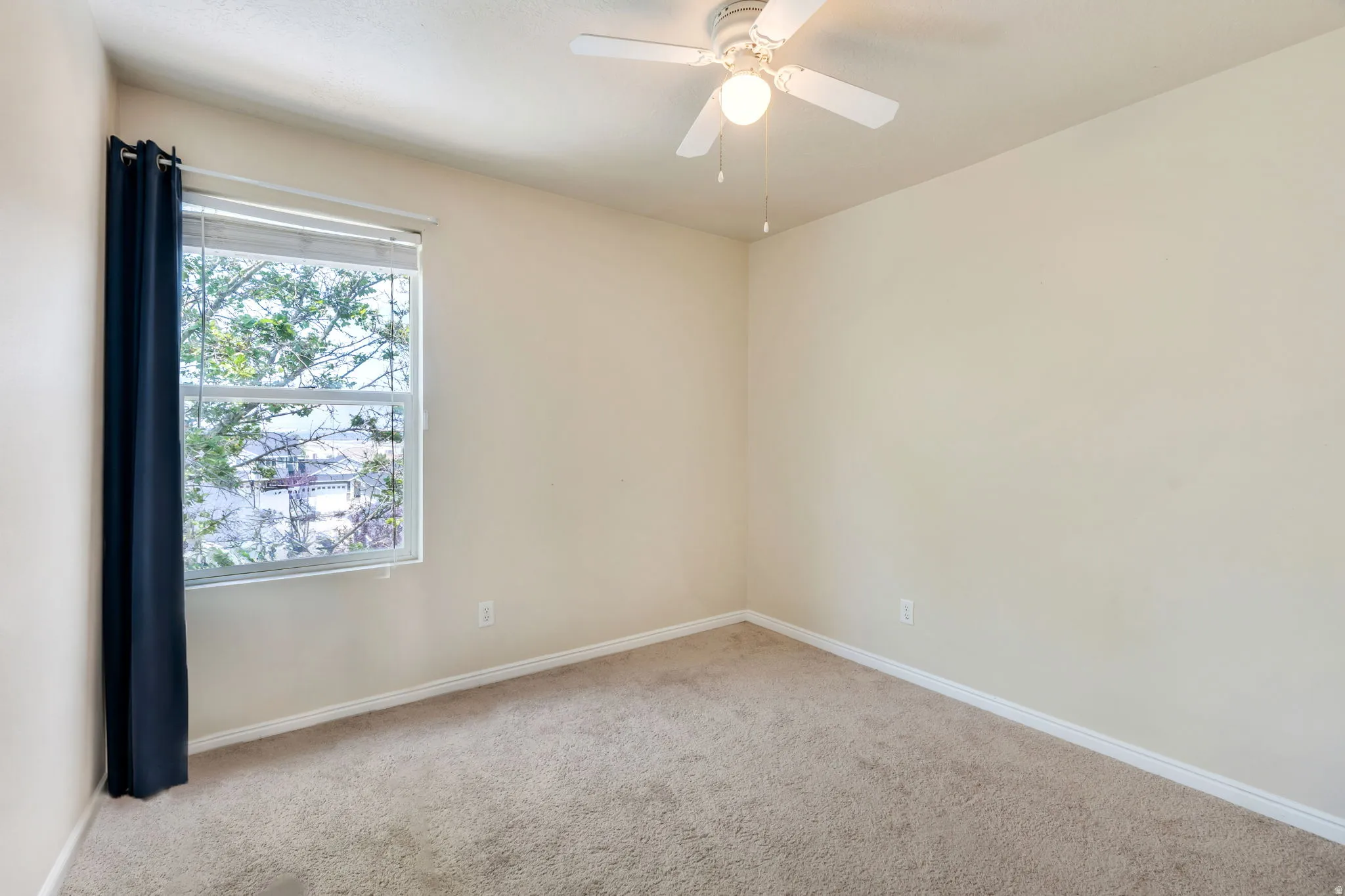 Bright room featuring a window with exterior tree scenery, a ceiling fan with integrated lighting, and neutral-toned wall-to-wall carpeting