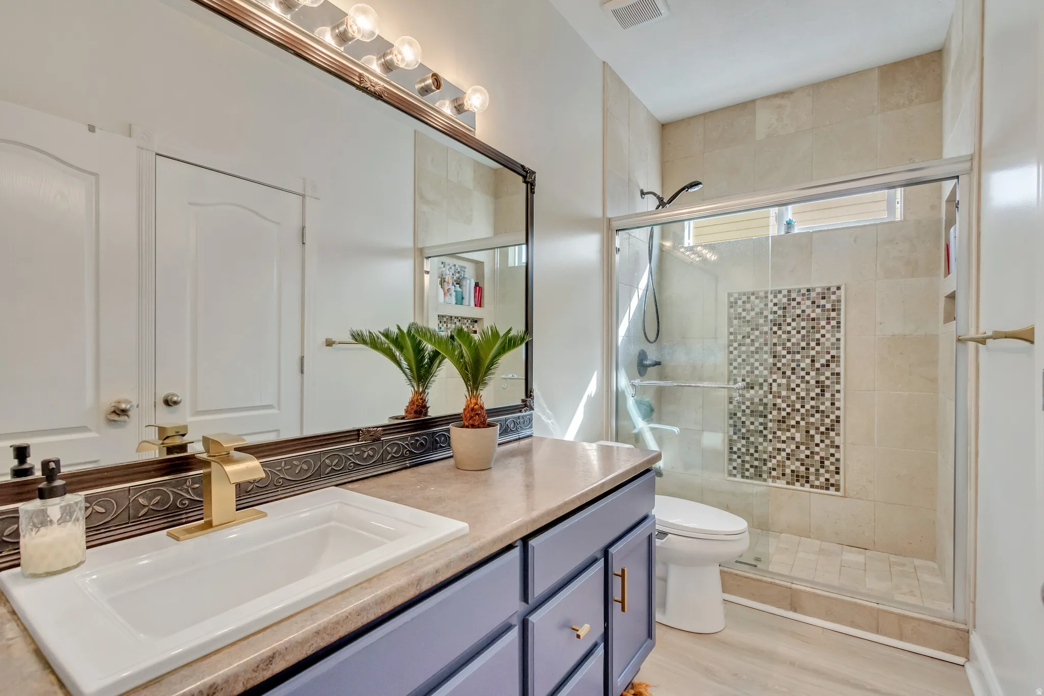 Bathroom featuring a single vanity with a rectangular sink and gold-tone faucet, a large mirror with integrated vanity lighting, a glass-enclosed shower with tile surround, and mosaic tile accent