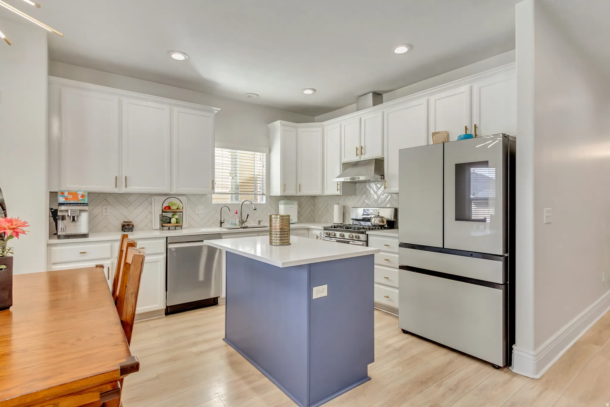 Bright kitchen featuring white shaker cabinetry, stainless steel appliances, a herringbone tile backsplash, a central island with a contrasting base, and light wood-finish flooring
