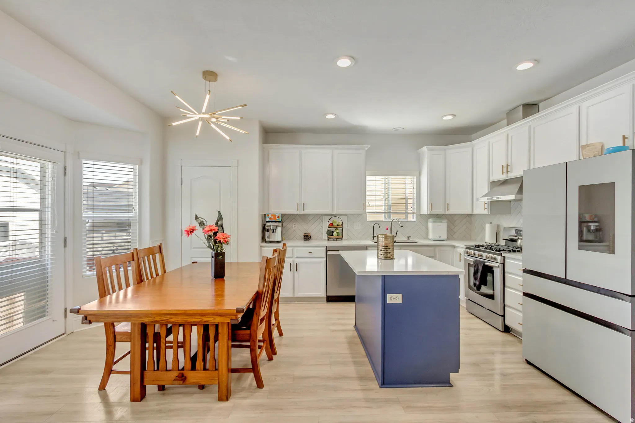Open-concept kitchen and dining area featuring light wood-finish flooring, a contemporary dining chandelier, recessed lighting, white cabinetry, a blue kitchen island with a beautiful quartz countertop, and stainless steel appliances less than 2 years old