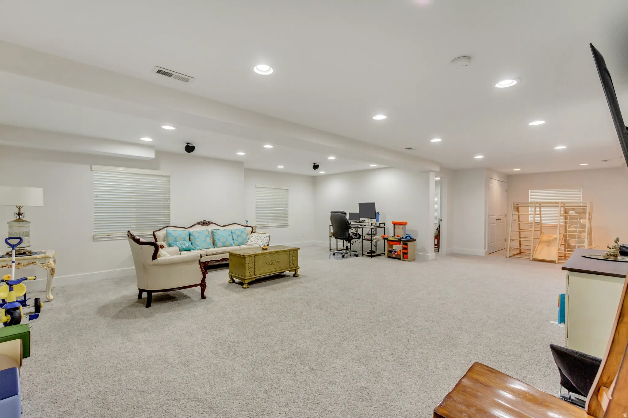Spacious lower-level room featuring light gray carpeting, recessed ceiling lighting, and multiple windows with horizontal blinds