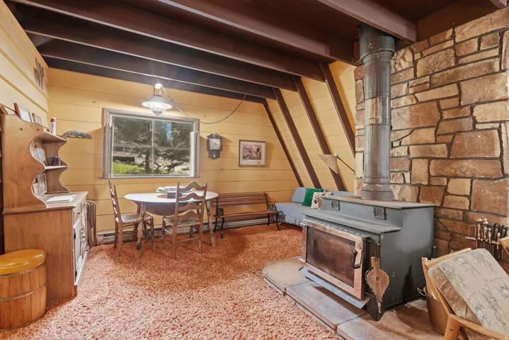 A-frame interior featuring a stone accent wall with a built-in wood-burning stove, exposed wood ceiling beams, and horizontal wood paneling