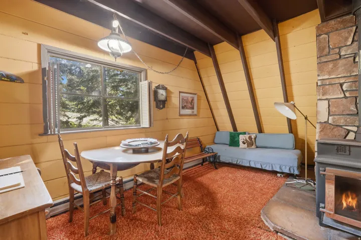 Living area featuring a stone-clad fireplace with an attached wood-burning stove, sloped wood-paneled walls, and exposed ceiling beams