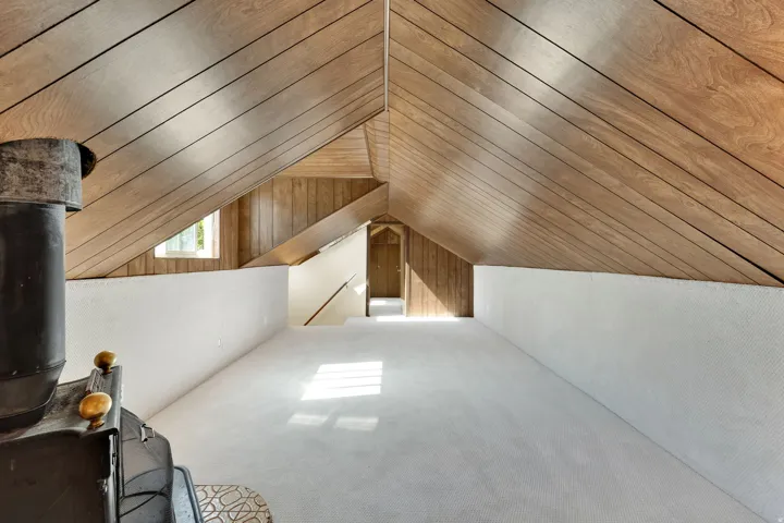 Spacious loft area featuring sloped wood-paneled ceilings, light-toned carpeting, and a freestanding wood-burning stove