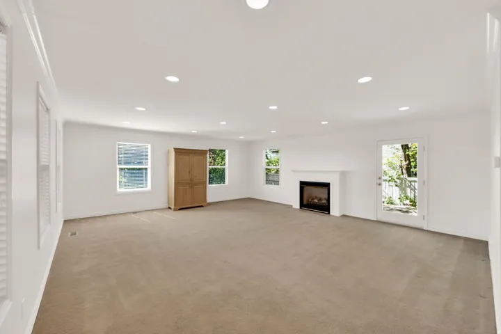 Spacious room featuring neutral carpet flooring, recessed lighting, multiple windows, a white fireplace with a dark insert, and a glass-paned exterior door