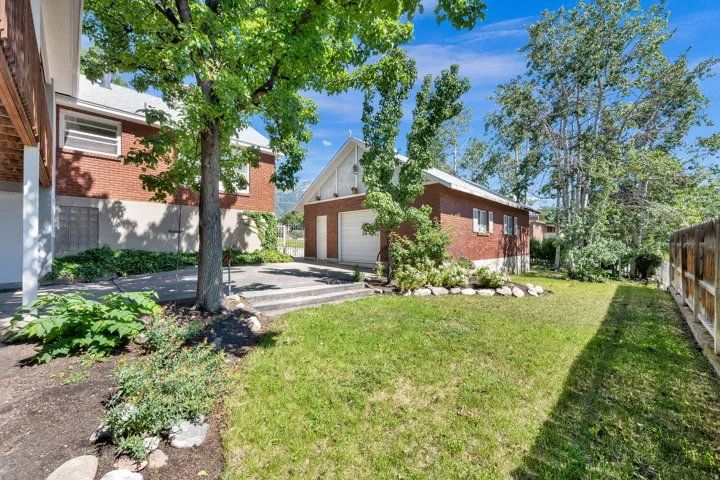 Expansive green lawn area featuring mature trees, a concrete patio, and a detached two-car garage with brick siding and an A-frame roofline