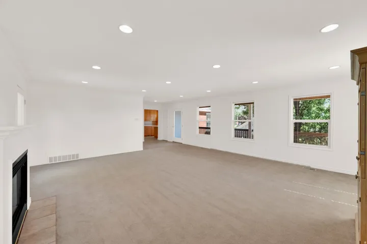 Spacious open-plan room featuring neutral carpeting, a white mantel fireplace, recessed lighting, and multiple windows with blinds