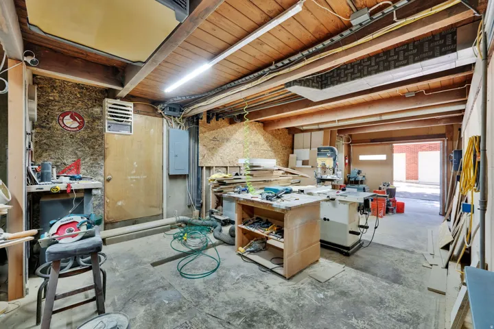Spacious workshop featuring exposed wood beam ceiling, concrete flooring, fluorescent overhead lighting, and an overhead garage door