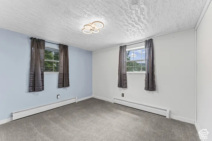 Carpeted room featuring two windows with curtain rods, baseboard heating, a modern ceiling light fixture, and a textured white ceiling