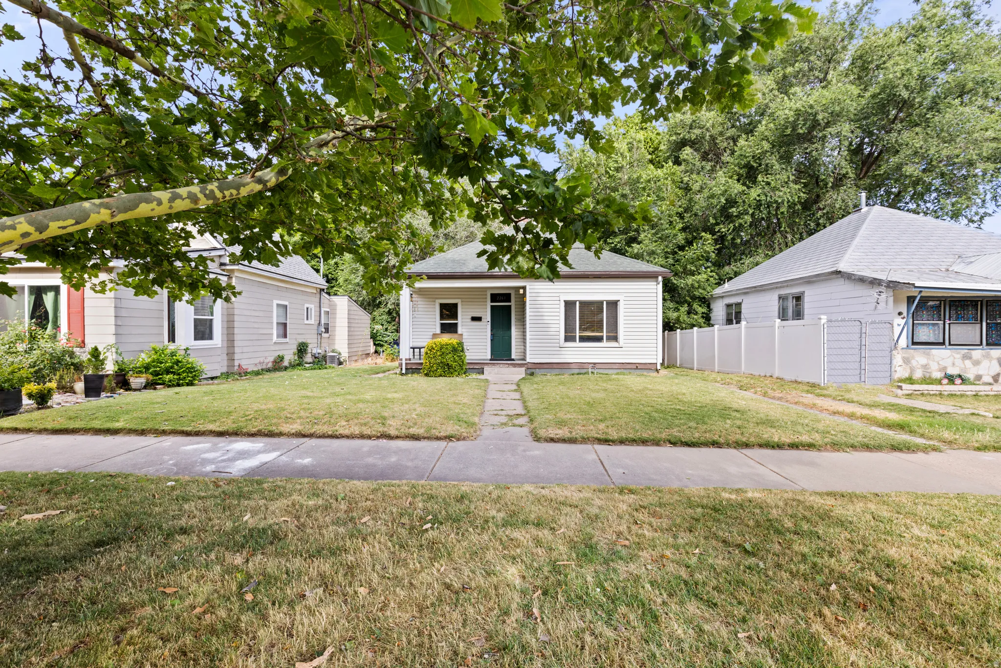 White siding exterior with a dark green front door, front yard with mature trees, and a concrete pathway leading to the entrance