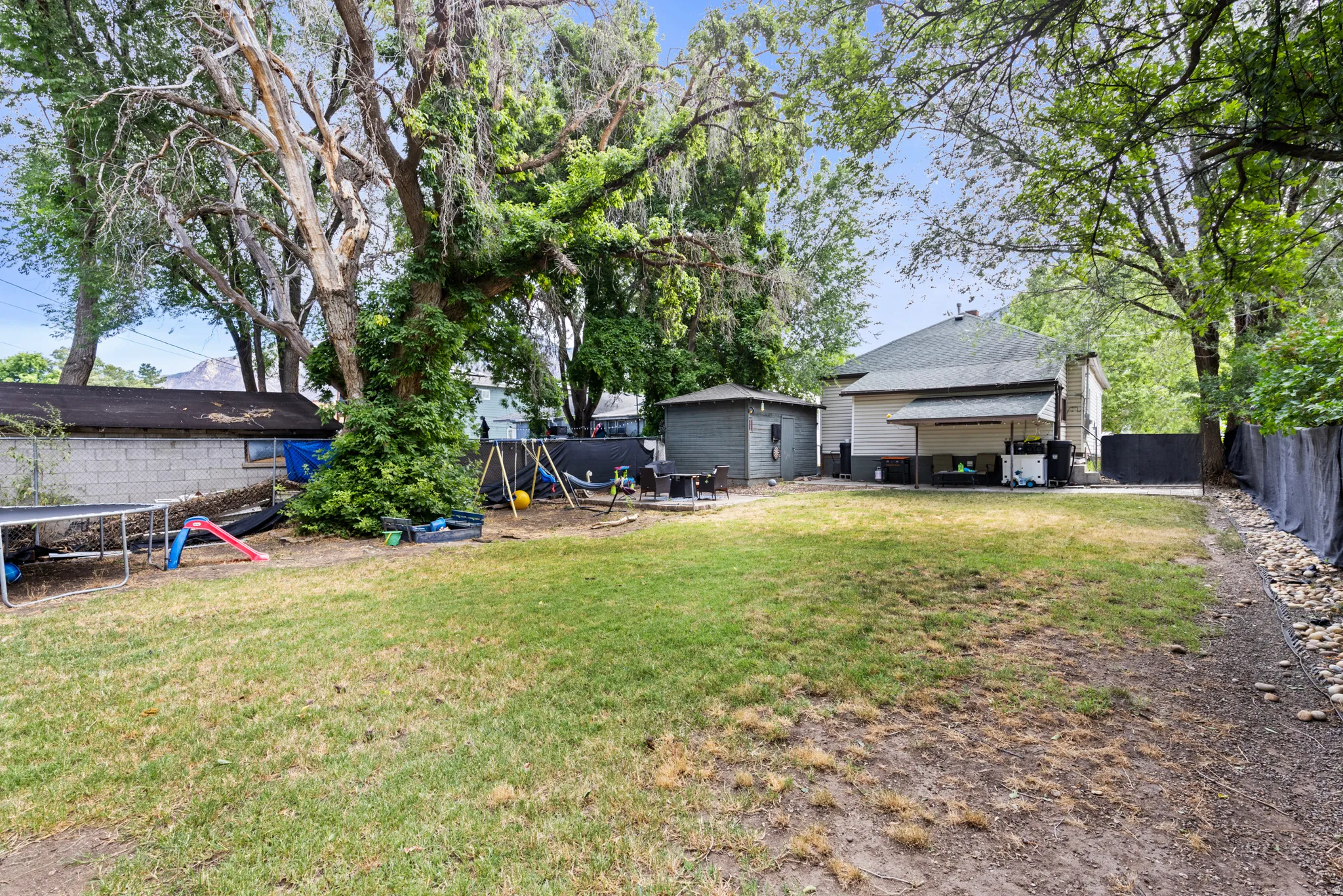 Expansive grassy yard area featuring mature trees, a permanent storage shed, and a covered patio area on the rear of the property