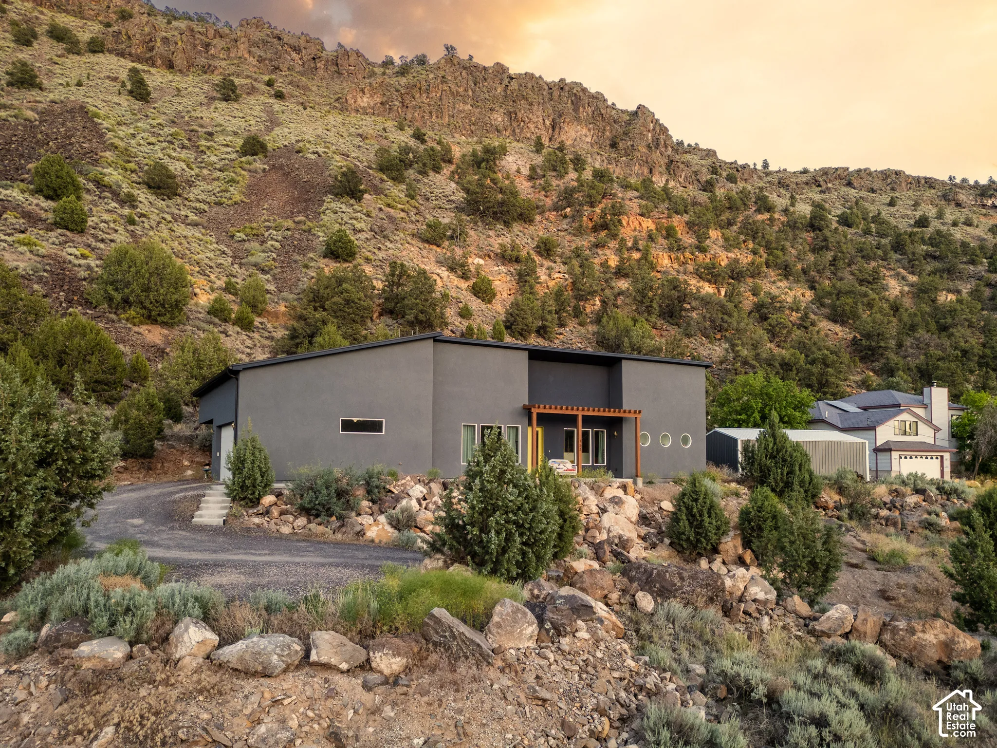 Contemporary grey exterior with a flat roof, featuring a wooden pergola over a patio, round accent windows, and a gravel driveway