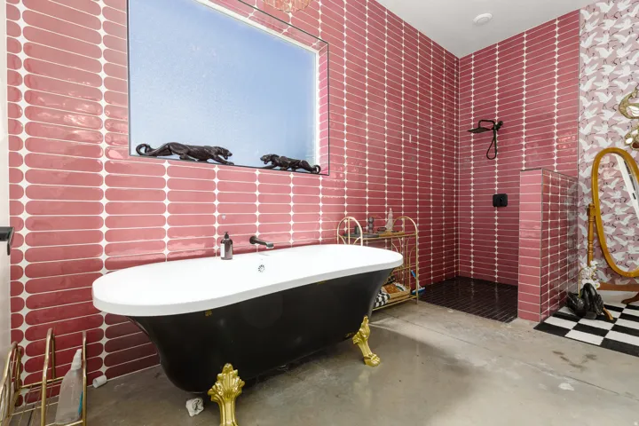 Freestanding clawfoot bathtub with gold-finish feet, walk-in shower with dark tile flooring, and pink elongated subway tile walls