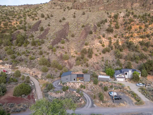 Modern gray exterior with dark trim and a contrasting wood-finish accent panel, set into a natural hillside landscape with a winding gravel driveway