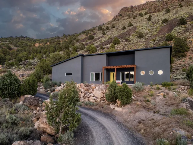 Contemporary exterior with a dark grey facade, a prominent yellow entry door, a wooden pergola, multiple circular windows, and a gravel driveway