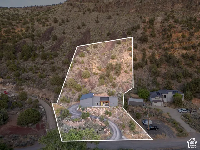 Contemporary hillside residence featuring a gray exterior, a dark-colored roof, a winding driveway, and surrounding natural terrain