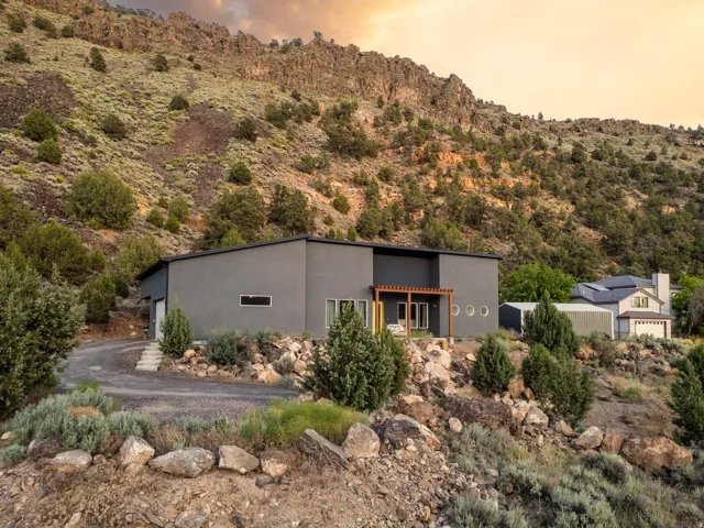 Contemporary grey exterior with a flat roof, featuring a wooden pergola over a patio, round accent windows, and a gravel driveway