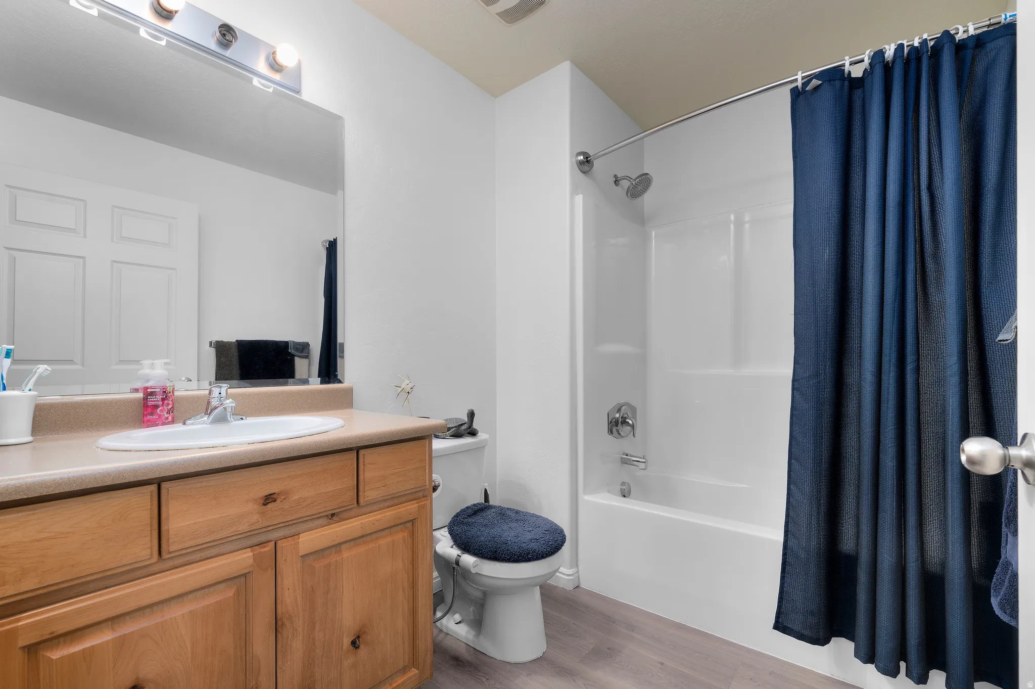 Bathroom featuring a light wood-finish vanity with an integrated sink, a large wall-mounted mirror, a three-light vanity bar, a white tub with shower, and wood-finish flooring