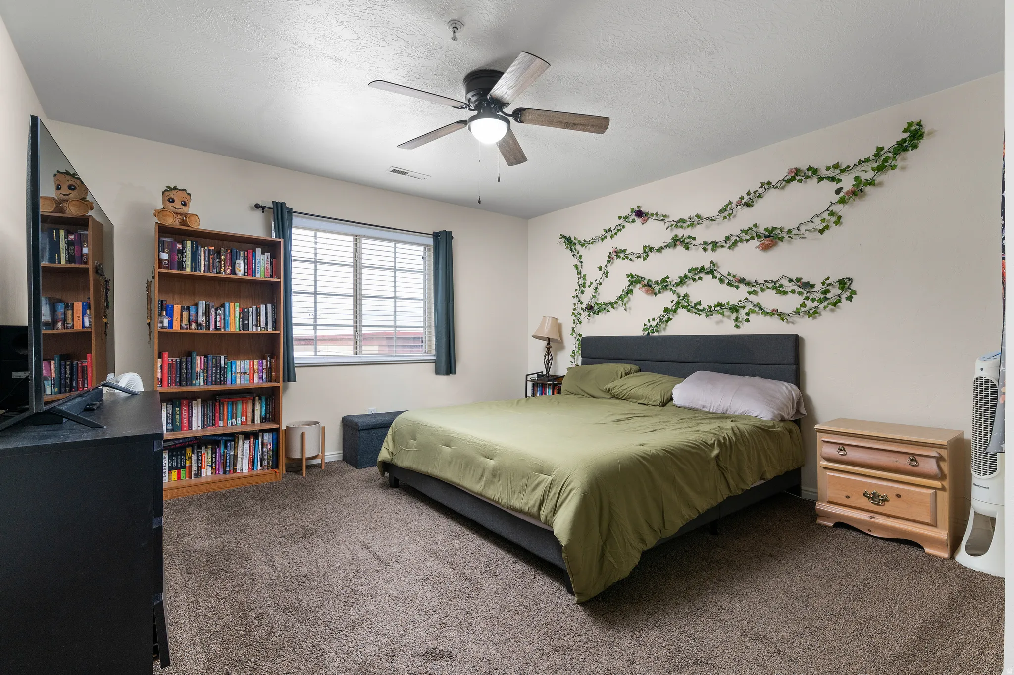 Carpeted bedroom featuring a ceiling fan with integrated lighting, a window with horizontal blinds, a built-in bookshelf, and light-toned walls