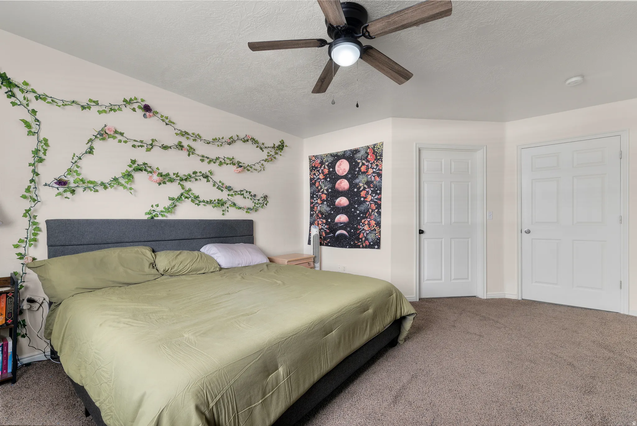 Carpeted room featuring a ceiling fan with integrated lighting, a substantial bed frame with an upholstered headboard, and two six-panel interior doors